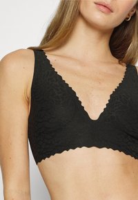 Nainen, jolla on yllään musta pitsinen bralette, jossa on aaltoilevat reunat ja teksturoitu kukkakuosi, joka näkyy ylävartalosta ja osasta hänen kasvojaan.