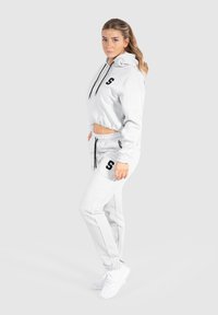 Hellgrauer, verkürzter Hoodie mit schwarzem Logo, kombiniert mit passenden, schmal zulaufenden Jogginghosen mit seitlichen Streifen und elastischen Bündchen, getragen mit weißen Sneakers.