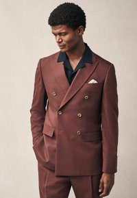Traje burdeos de doble botonadura con un acabado texturizado, que presenta botones de latón y una camisa negra de cuello debajo. Acento de pañuelo blanco en el bolsillo.