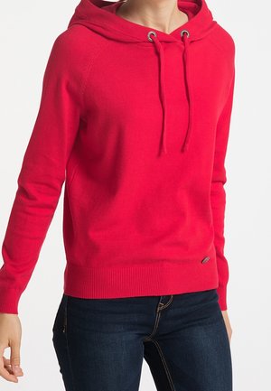 Frau trägt einen leuchtend roten Kapuzenpullover mit Kordelzug in Kombination mit dunkelblauen Jeans vor weißem Hintergrund.