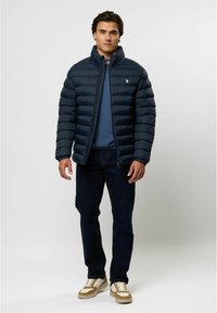 Marineblauwe gewatteerde jas met horizontale stiksels, hoge kraag en rits. Draag over een blauwe trui, gecombineerd met donkere spijkerbroek en sneakers.