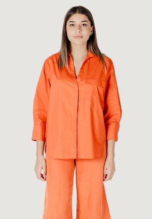 Camicia arancione con colletto, maniche lunghe e tasca sul petto. La camicia è realizzata in tessuto morbido, abbinata a pantaloni arancioni coordinati.