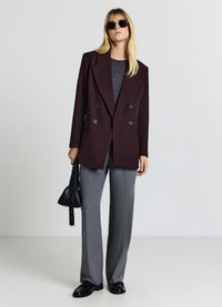 Double-breasted burgundy blazer with large black buttons, paired with gray patterned trousers. Model carries a small black bag.