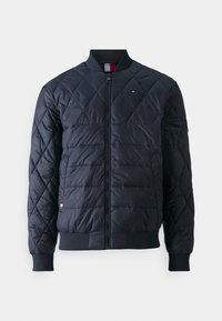 Svart quiltad bomberjacka med framdragkedja, ribbad krage och muddar, samt en liten röd, vit och blå logotyp på bröstet och fållen.