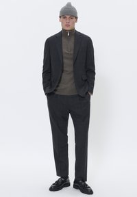 Ung mand iført en grå beanie, mørkternet jakkesæt med matchende bukser, brun zip-up sweater og sorte loafers, står med hænderne i lommerne.