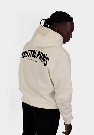 Crème hoodie met zwarte grafische tekst "CRYSTAL PARIS" en "fight for freedom" op de achterkant. Zachte textuur, ontspannen design en grote capuchon.