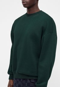 Homme portant un sweat-shirt à col rond vert foncé et un pantalon à carreaux foncé sur un fond clair uni.