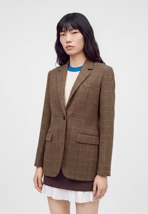 Jeune femme aux longs cheveux noirs portant un blazer à carreaux marron sur une jupe à plusieurs couches et un haut blanc, debout devant un fond uni.