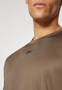 T-shirt de sport marron en tissu lisse, avec un col rond et un petit logo noir sur la poitrine.