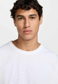 Camiseta blanca de algodón con cuello redondo y sutil logotipo de Nike en relieve en el lado izquierdo del pecho. La textura parece suave y ligera.