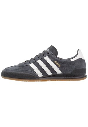 Zapatilla Adidas de ante gris oscuro con rayas blancas, suela de goma y la palabra "JEANS" en letras doradas en el lateral.