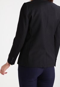 Blazer negro ajustado con mangas largas y puños abotonados. Presenta un diseño estructurado y una tela suave y texturizada.