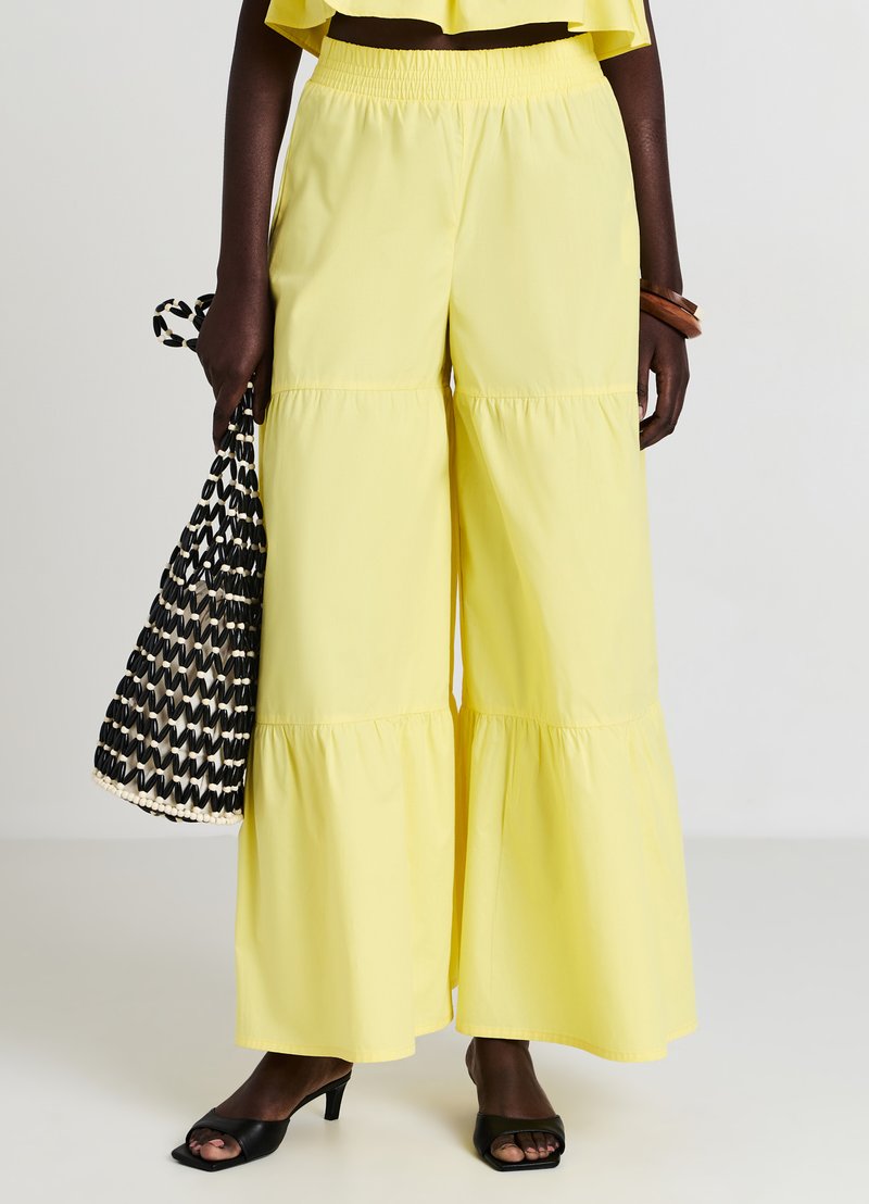 Gelbe weit geschnittene Pants mit einem gestuften Rüschensaum und einem elastischen Bund. Kombiniert mit einer schwarz-weißen gewobenen Tasche und schwarzen Blockabsatzsandalen.
