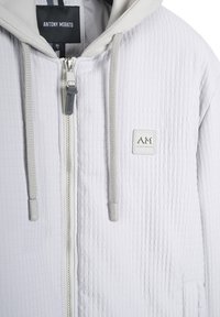 Svijetlosiva teksturirana zip-up hoodie s vezicama, oznaka branda na prsima i oznaka "Antony Morato" unutar ovratnika.
