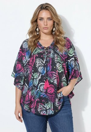 Woman with wavy blonde hair wearing a loose black blouse with colorful butterfly prints and blue jeans, standing with one hand in pocket.