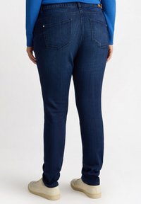 Jeans ajustados de color azul oscuro con cinco bolsillos y una textura suave, combinados con zapatillas de color claro, vistos desde atrás.