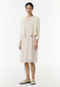 Lichtroze geplooide jurk met een sleutelgat halslijn, gecombineerd met een crèmekleurige cropped cardigan en beige instapschoenen met een lage zwarte zool.