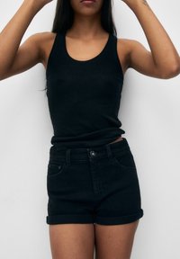 Débardeur noir associé à un short en jean noir, avec une coupe ajustée, des revers roulés et une texture lisse. Le modèle pose devant un fond neutre.