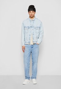 Giacca di jeans azzurro chiaro con tasche sul petto, t-shirt crema sotto, jeans azzurro chiaro e berretto nero. Scarpe da ginnastica bianche completano l'ensemble.