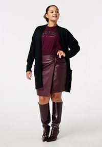 Vrouw met oorbellen in de vorm van een ring, zwarte open cardigan, bordeauxrode "LOVER" t-shirt, bordeauxrode neplederen rok en bijpassende kniehoge laarzen.