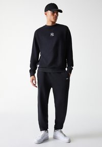 Schwarzer Sweatshirt mit weißem Logo auf der Brust, kombiniert mit schwarzen Jogginghosen. Das Modell trägt eine schwarze Mütze und weiße Sneaker. Glatter, lässiger Stoff.