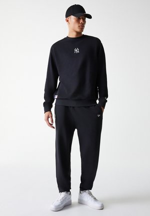 Black sweatshirt with white logo on chest, paired with black jogger pants. Model wears a black cap and white sneakers. Smooth, casual fabric.