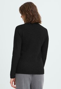 Donna con capelli castani di lunghezza media che indossa un maglione nero aderente e pantaloni grigi, girata di spalle su uno sfondo neutro.