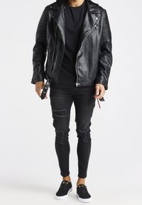 Giacca da motociclista nera in pelle con design a coste sulle spalle, abbinata a jeans skinny neri strappati e sneakers nere con suole bianche.