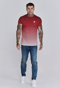 Hombre con tatuajes que lleva una camiseta de degradado rojo a blanco, jeans azules y zapatillas blancas, de pie frente a un fondo liso claro.