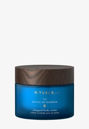 Blå krukke af Rituals The Ritual of Hammam pisket body cream med låg med mørkt trætekstur på en ensfarvet hvid baggrund.