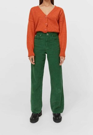Personne portant un cardigan orange boutonné, un pantalon en velours côtelé vert taille haute et des chaussures à plateforme noires, se tenant devant un fond uni.