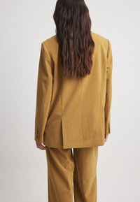 Traje ajustado de color mostaza con hombros estructurados, corte recto, una abertura en la parte posterior y puños abotonados en las mangas.