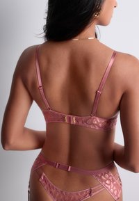 Femme portant un ensemble de lingerie en dentelle rose avec des bretelles ajustables, vue de dos sur un fond uni clair.