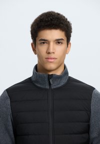 Chaleco de plumas negro con mangas tipo suéter en gris, cuello alto y cremallera frontal; presenta una textura acolchada y un tejido en contraste.