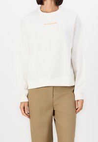 Sudadera de forro blanco con mangas largas, cuello redondo, logo naranja "Marc O'Polo Denim" en el pecho, combinada con pantalones beiges.