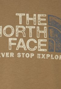 Logo The North Face s částečně viditelným sloganem "Nikdy nepřestávej objevovat" na hnědém textilním pozadí.