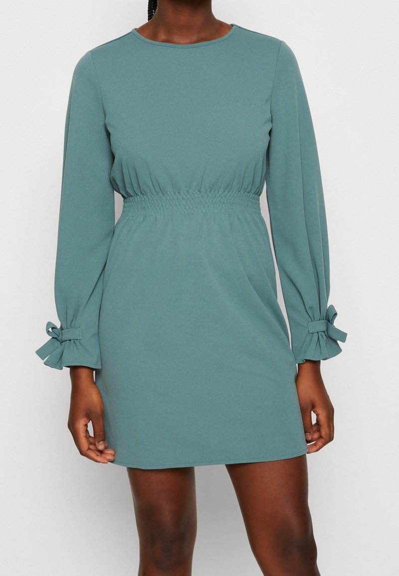 Robe à manches longues bleu sarcelle avec taille froncée et poignets noués, portée par une personne sur un fond clair uni.