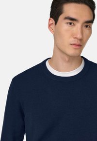 Pull en maille bleu marine à col rond, avec une texture douce et une coupe légèrement ajustée, porté sur une chemise blanche.
