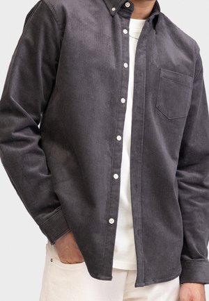 Homme portant une chemise en velours côtelé gris foncé boutonnée sur un t-shirt blanc, avec la main dans la poche d’un pantalon couleur crème.