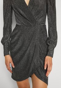 Femme portant une robe portefeuille métallisée gris foncé à manches longues, col en V et ourlet asymétrique, debout, les mains détendues le long du corps.