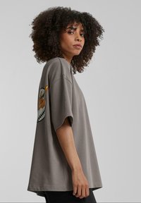 Camiseta de gran tamaño en gris, hecha de algodón suave, con mangas cortas, un cuello redondeado y un gráfico temático de béisbol en el brazo izquierdo.