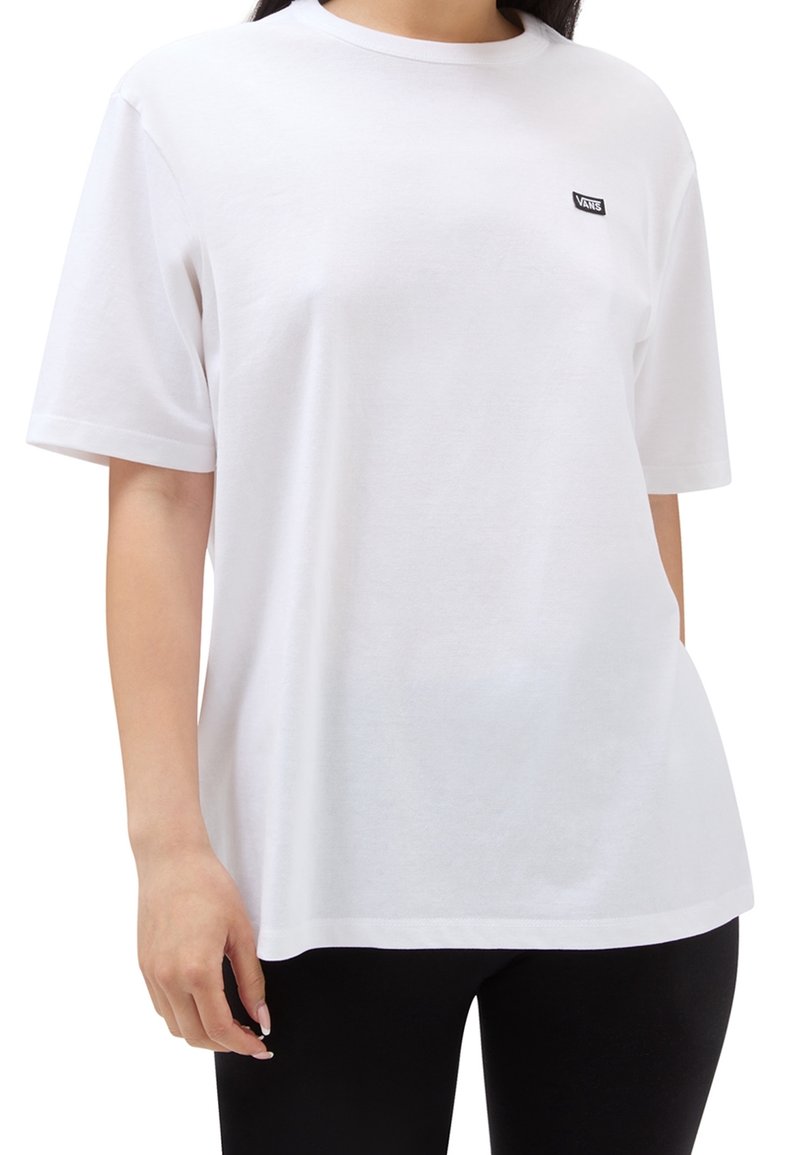 Femme portant un t-shirt blanc uni Vans avec un petit logo sur la poitrine gauche, associé à un pantalon noir.