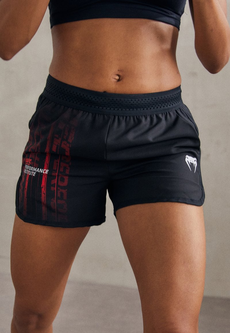 Personne tonique portant un short noir UFC Performance Institute et un haut de sport noir, debout devant un fond uni.