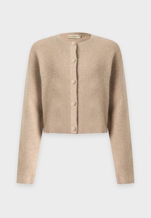 Femme aux cheveux noirs porte un cardigan beige boutonné, un col roulé foncé, un short plissé rose clair, des chaussettes noires montant jusqu'aux genoux et des bottes en cuir noires.