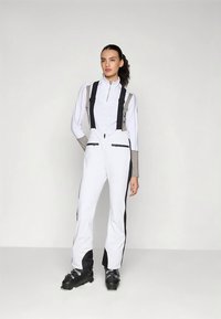 Vrouw in een witte en beige skikleding met zwarte bretels en zwarte skischoenen staand tegen een effen witte achtergrond.