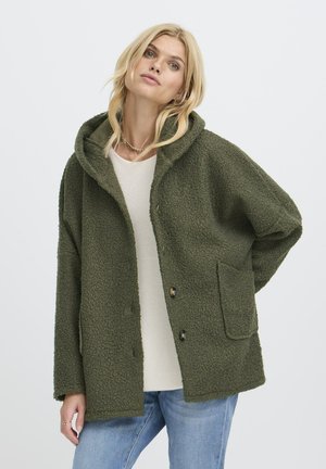 Lyshåret kvinde iført grøn tekstureret hættejakke over hvid top og blå jeans, står med den ene hånd bag ryggen mod en ensfarvet baggrund.