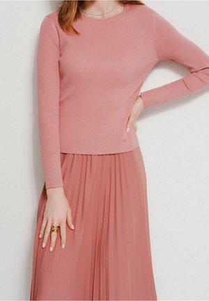 Femme portant un top rose à manches longues côtelé et une jupe plissée rose, avec des bagues en or à la main gauche et un collier délicat.