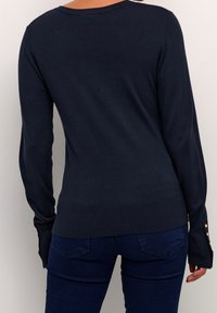 Námořnický modrý pletený svetr s dlouhými rukávy, žebrovaným lemem a knoflíkovými akcenty na manžetách rukávů. Hladká textura, jednoduchý design a přiléhavý styl.