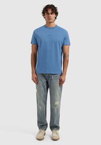Lichtblauwe katoenen t-shirt met een ronde hals en korte mouwen, voorzien van een bedrukte zwarte logo op de borst. Gedragen met versleten denim jeans.