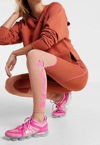 Sportowy strój składający się z pomarańczowej bluzy, cielistych legginsów z różowymi akcentami oraz różowych sneakersów z przezroczystą podeszwą.
